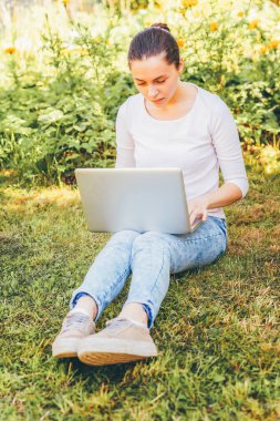 Mobil Ofis. Serbest iş konsepti. Şehir parkında yeşil çimlerde oturan genç bir kadın dizüstü bilgisayarla çalışıyor. Yaşam tarzı otantik, açık havada okuyan samimi öğrenci kız.