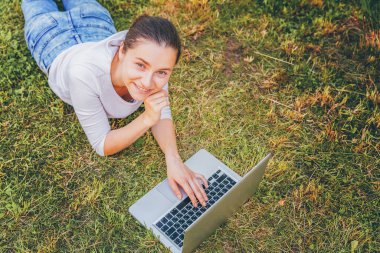 Mobil Ofis. Serbest iş konsepti. Şehir parkında çimlerin üzerinde yatan genç bir kadın dizüstü bilgisayarla çalışıyor. Yaşam tarzı otantik, açık havada okuyan samimi öğrenci kız..