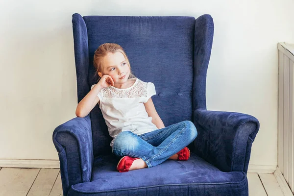 Stay Home Stay Safe. Sweet little girl in jeans and white T-shirt sitting on modern cozy blue chair relaxing in white bright living room at home indoors. Childhood schoolchildren youth relax concept