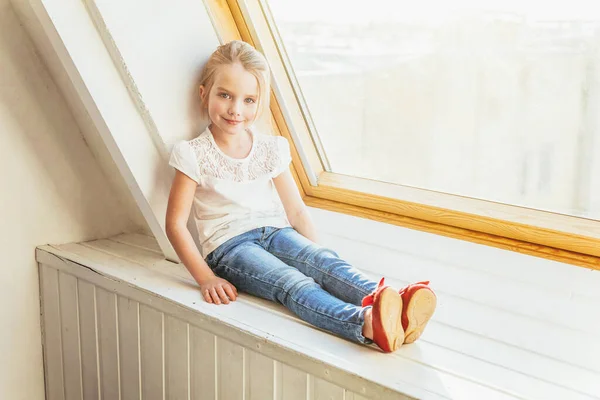 Stay Home Stay Safe. Little cute sweet smiling girl in jeans and white T-shirt sitting on window sill in bright light living room at home indoors. Childhood schoolchildren youth relax concept