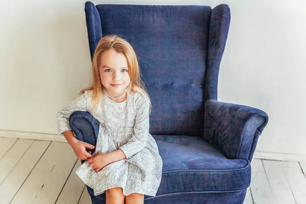 Stay Home Stay Safe. Sweet little girl in white dress sitting on modern cozy blue chair relaxing in white bright living room at home indoors. Childhood schoolchildren youth relax concept