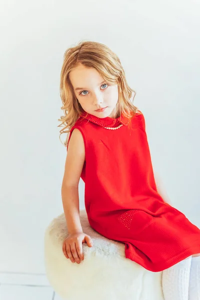 Stay Home Stay Safe. Sweet little girl in red dress sitting on chair against white wall at home, relaxing in white bright living room indoors. Childhood schoolchildren youth relax concept