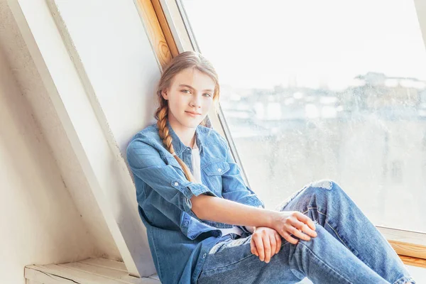 Stay Home Stay Safe. Young cute teenage girl in jeans, denim jacket and white T-shirt sitting on window sill in bright light living room at home indoors and thinking. Social distance jomo concept