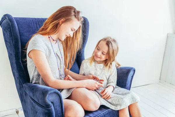 Stay Home Stay Safe. Two happy kids sitting on cozy blue chair relaxing playing in white living room indoors. Little girl playing with teenage girl showing her love care. Sisters having fun at home