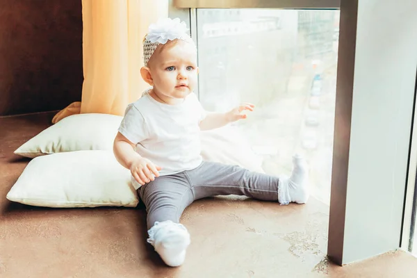 Blijf veilig thuis. Klein schattig baby meisje zit in de buurt van groot raam in helder licht woonkamer thuis binnen. Het begrip "gevoeligheid voor moederschap tijdens de kindertijd". — Stockfoto