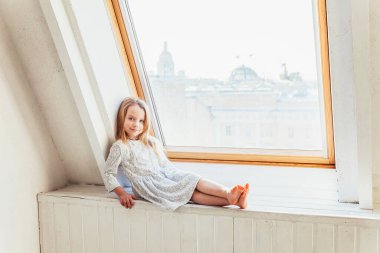 Evde kal. Kendine dikkat et. Beyaz elbiseli, sevimli, tatlı bir kız. Evdeki parlak ışıklı oturma odasında pencere pervazında oturuyor. Çocukluk çocukları gençlik konsepti