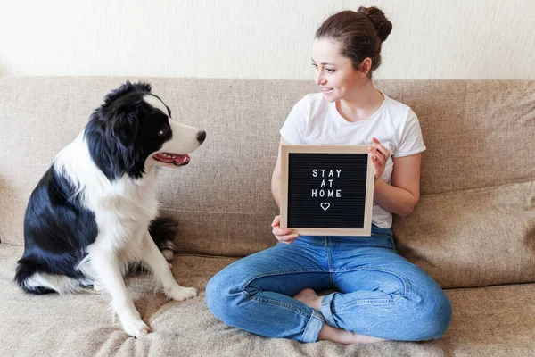 Stay Home Stay Safe. Smiling young woman playing with cute puppy dog border collie on sofa at home indoors. Girl with letter board inscription STAY AT HOME. Pet care animal life quarantine concept