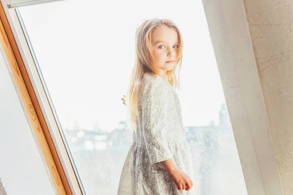 Stay Home Stay Safe. Little cute sweet smiling girl in white dress standing on window sill in bright light living room at home indoors. Childhood schoolchildren youth relax concept