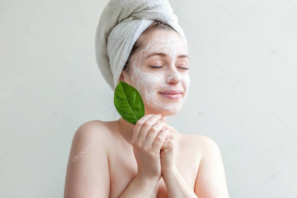 Minimal beauty portrait woman girl in towel on head applying white nourishing mask or creme on face, green leaf in hand isolated white background. Skincare cleansing eco organic cosmetic spa concept