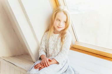 Evde kal. Kendine dikkat et. Beyaz elbiseli, sevimli, tatlı bir kız. Evdeki parlak ışıklı oturma odasında pencere pervazında oturuyor. Çocukluk çocukları gençlik konsepti.