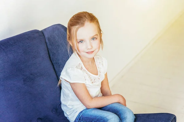 Restez à la maison Restez en sécurité. Douce petite fille en jean et T-shirt blanc assis sur une chaise bleue confortable moderne relaxant dans le salon lumineux blanc à la maison à l'intérieur. Enfance écoliers jeunesse relax concept . — Photo