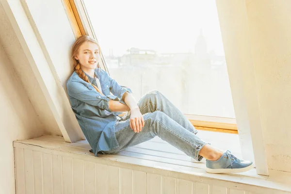 Stanna hemma och var säker. Ung söt tonåring flicka i jeans, jeans jacka och vit T-shirt sitter på fönsterbräda i ljust vardagsrum hemma och tänker. Begreppet socialt avstånd. — Stockfoto