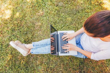 Serbest iş kavramı. Dizüstü pc bilgisayar üzerinde çalışan Şehir Parkı yeşil çim Çimenlikte oturan genç kadın. Yaşam tarzı otantik samimi öğrenci kız açık havada okuyor. Mobil ofis