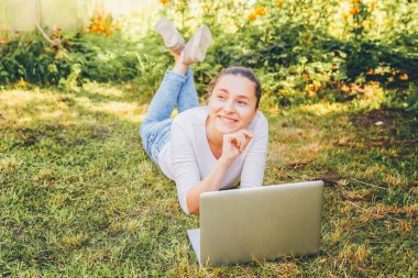 Mobil Ofis. Serbest iş konsepti. Şehir parkında çimlerin üzerinde yatan genç bir kadın dizüstü bilgisayarla çalışıyor. Yaşam tarzı otantik, açık havada okuyan samimi öğrenci kız..