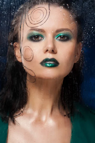 Mooi model meisje staat achter een glas met regendruppels. — Stockfoto
