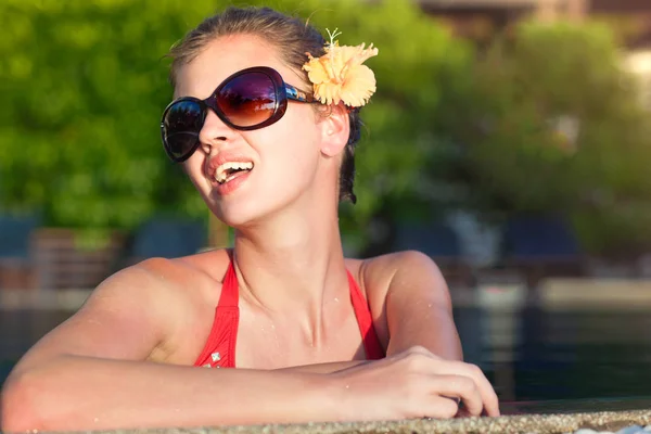 Hermosa mujer joven en gafas de sol con flor en el pelo sonriendo en la piscina de lujo —  Fotos de Stock