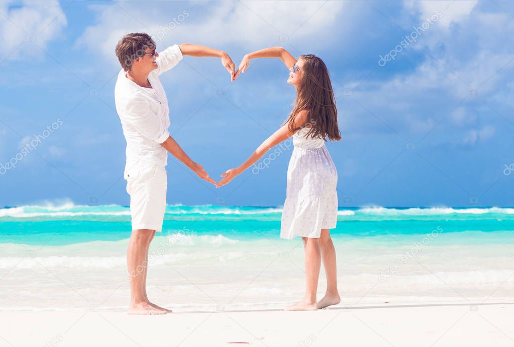 young happy couple in white making heart shape on tropical beach. honeymoon