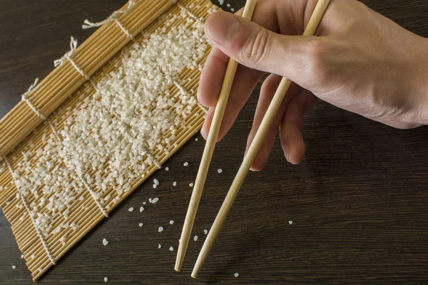 Palillos en la mano en el fondo de una estera de sushi y arroz disperso — Foto de Stock