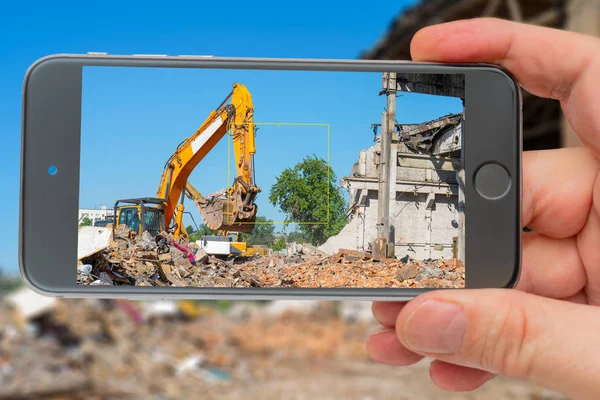 Destruction of old building. Yellow excavator on ruins. Photo smartphone. Smartphone in hand. Digger on screen.