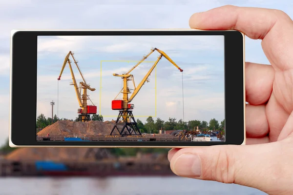 Construction crane on the smartphone screen. Cranes operate in river port. Cargo port on river.