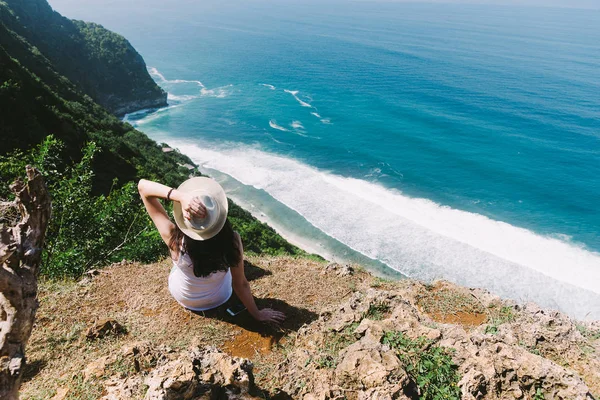 En flicka sitter på kanten av en klippa och beundra havet. En flicka sitter på kanten av en klippa. Fantastisk utsikt. Resa på det ö av Bali.Indonesia. — Stockfoto