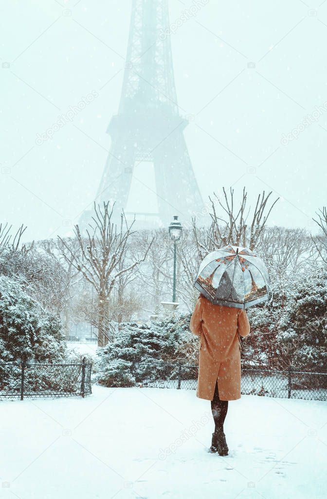 A young woman in a beige coat walks under an umbrella in a snowy winter Paris against the backdrop of the Eiffel Tower. Romantic brunette girl is walking in the background of falling snow in Paris. Snowy paris