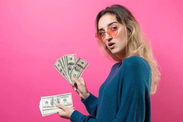 ピンクの背景の上に手にアメリカドルのお金を持つ眼鏡の青いセーターの若い美しい女性の閉鎖 喜びの感情やジェスチャーを表現する — ストック写真