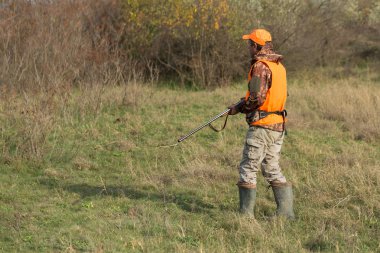 Elinde silahı olan bir avcı ve yansıtıcı turuncu yelekli bir av köpeği bozkırda bir sülün avlıyor..