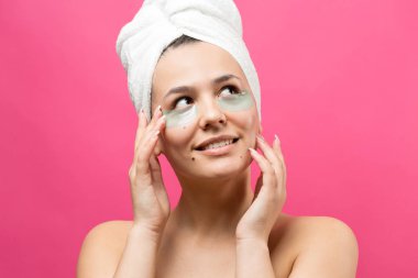 Kafasında beyaz havluyla gezen genç bir kız gözlerinin altına kolajen jel bantlar takıyor. Göz teması altında maske. 