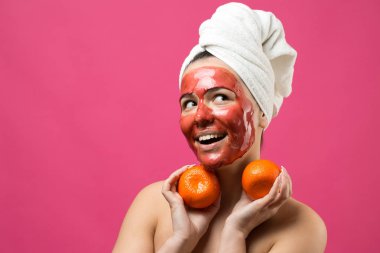 Kafasında kırmızı besleyici maske olan beyaz havluyla güzel bir kadın portresi. Cilt temizleme organik kozmetik rahatlama konsepti. Sırtı turuncu bir mandalina tutan bir kız..