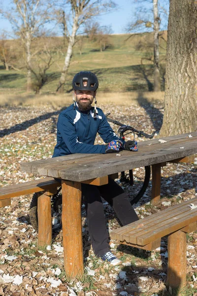 エアサスフォークがオフロードに乗っている現代のカーボンハードテールバイクのパンツやフリースジャケットでサイクリスト 男は秋の公園のベンチで休んでいる — ストック写真