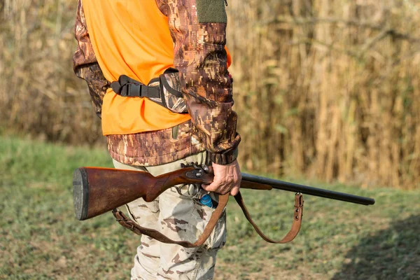 A man with a gun in his hands and an orange vest on a pheasant hunt in a wooded area in cloudy weather. Hunter with dogs in search of game.