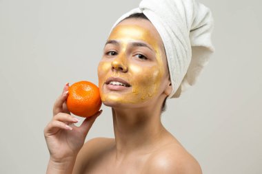Yüzünde altın besleyici maske olan beyaz havluyla kadın portresi. Cilt temizleme organik kozmetik rahatlama konsepti. Sırtı turuncu bir mandalina tutan bir kız..