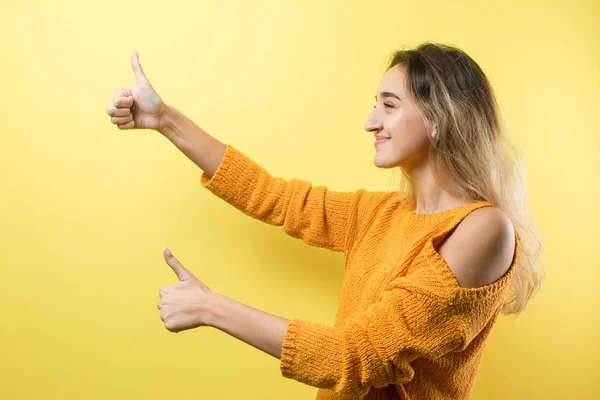 Happy young caucasian female in an orange sweater making thumb up sign and smiling. Good job and respect.