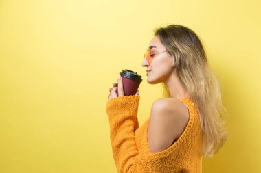 Gözlüklü, turuncu kazaklı, sarı arka planda kahve içen çekici bir kadın.