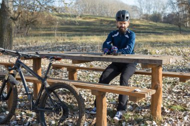 Pantolon ve yün ceketli bir bisikletçi modern karbon donanımlı bir motorsikletle off-road 'a biniyor. Adam sonbahar parkında bir bankta dinleniyor..