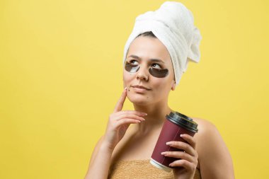 Kafasında beyaz havluyla gezen genç bir kız gözlerinin altına kolajen jel bantlar takıyor. Göz tedavisiyle maskelenmiş. Kız elinde bir bardak kahve tutuyor..