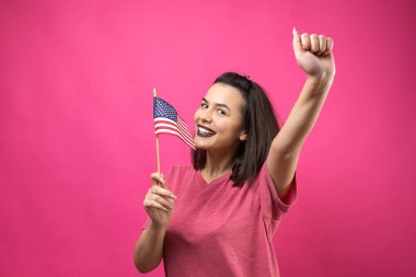 Stüdyonun pembe arka planında Amerikan bayrağı tutan mutlu genç kadın.