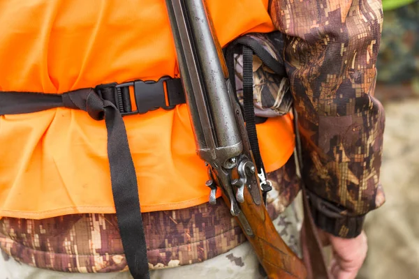 A man with a gun in his hands and an orange vest on a pheasant hunt in a wooded area in cloudy weather. Hunter with dogs in search of game.