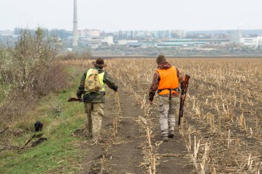 Elinde silah ve turuncu yelek olan bir adam bulutlu bir havada ormanlık alanda sülün avında. Oyun arayan köpeklerle avcılar.