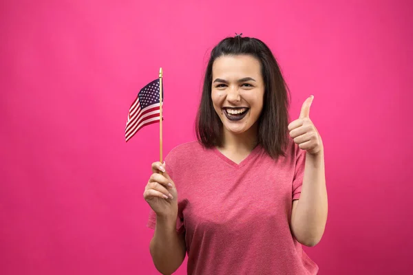 快乐的年轻女子拿着美国国旗面对着粉红的工作室背景 — 图库照片