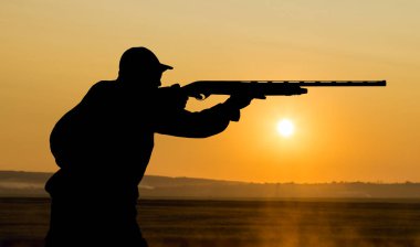 Elinde silah ve turuncu yelek olan bir adam bulutlu bir havada ormanlık alanda sülün avında. Avcı köpeklerle av peşinde.