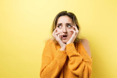 Sarı kazaklı, beyaz tişörtlü, uzun saçlı, yüzünde şaşırmış bir ifade olan bej arka planda yaşayan genç bir kadın.