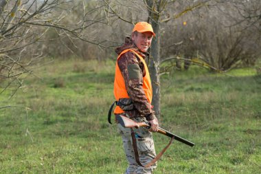 Elinde silah ve turuncu yelek olan bir adam bulutlu bir havada ormanlık alanda sülün avında. Avcı köpeklerle av peşinde.