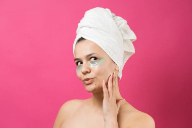 Kafasında beyaz havluyla gezen genç bir kız gözlerinin altına kolajen jel bantlar takıyor. Göz teması altında maske.