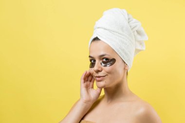 Kafasında beyaz havluyla gezen genç bir kız gözlerinin altına kolajen jel bantlar takıyor. Göz teması altında maske.