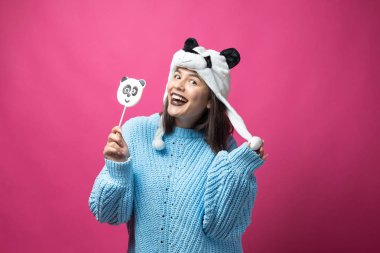 Elinde lezzetli bir panda lolipopu ve pembe bir arka planda şapkası olan komik bir genç kız..
