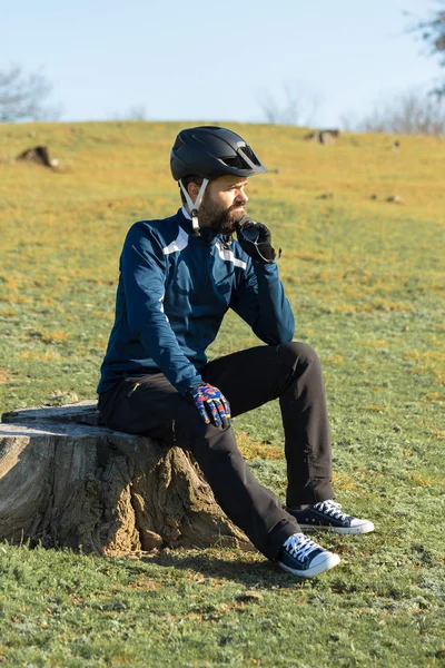 エアサスフォークがオフロードに乗っている現代のカーボンハードテールバイクのパンツやフリースジャケットでサイクリスト あの男は公園の切り株の上で休んでる遠くを見て — ストック写真