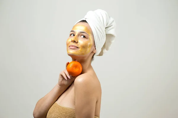 头戴白色毛巾 脸上戴金色营养面罩的女人的美丽画像 瘦身清洁生态有机化妆品温泉放松的概念 一个女孩背着橙子站在那里 — 图库照片