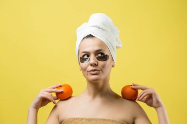 Yüzünde altın besleyici maske olan beyaz havluyla kadın portresi. Cilt temizleme organik kozmetik rahatlama konsepti. Sırtı turuncu bir mandalina tutan bir kız..